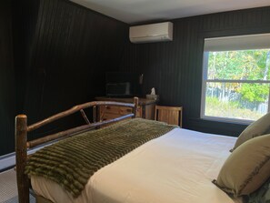 A bedroom with a wooden bed frame, a green blanket, a white mattress, a window with a view of trees, and an air conditioning unit on the wall. - Hatch's Entire Flock  - 5 unit compound (Cooperstown)