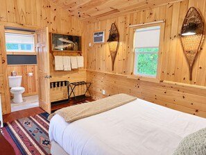 A wooden cabin bedroom with a bed, a window with a view, a bathroom with a toilet, and a shelf with towels. - Hatch's Coop - Awesome lake views, great studio cottage (Cooperstown)