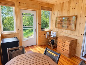 A wooden cabin interior with a dining table, chairs, a small kitchen area with a coffee maker, and a window overlooking a green landscape. - Hatch's Coop - Awesome lake views, great studio cottage (Cooperstown)