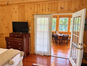 A wooden room with a television, a wooden dresser, a bed with a blanket, and a door leading to an outdoor area with a table and chairs. - Hatch's Coop - Awesome lake views, great studio cottage (Cooperstown)