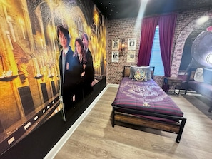 Een kamer met een bakstenen muur, een groot poster van een tovenaar en heks, een bed met een sierkussen met het Hogwarts-wapen, en een klein tafeltje met een lamp.