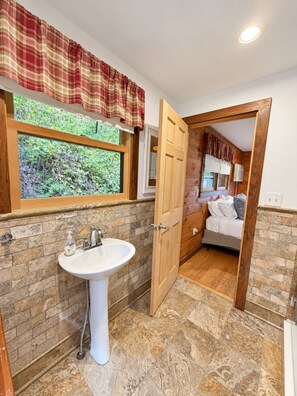 A bathroom with a white sink, a window with a view of greenery, and a wooden door leading to a bedroom. - Hatch's Roost -  Classic Otsego Camp, shared waterfront (Cooperstown)