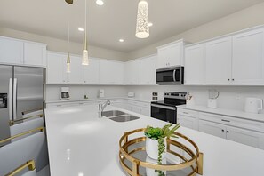 A modern kitchen with white cabinets, stainless steel appliances, and a central island with a gold-rimmed glass top. - Luxury 10br Resort Home w/ Pool & Movie Theater (Clermont)