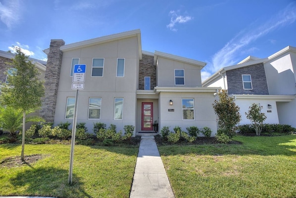 A modern two-story building with a red door, a wheelchair accessibility sign, and a well-maintained lawn. - Bookmark Villa 8BR @ Storey Lake w/ Pool (Kissimmee)