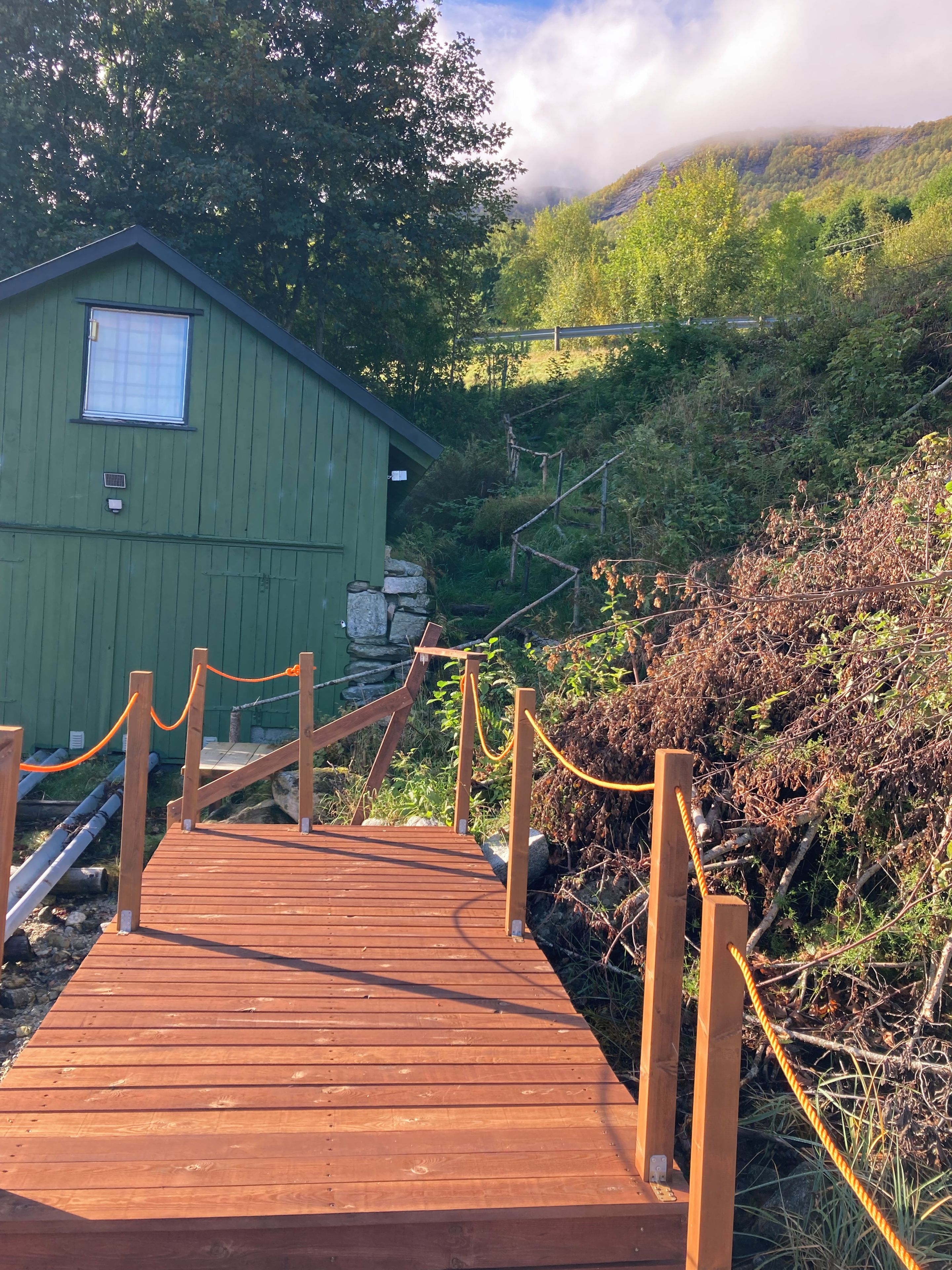 A wooden deck leading to a green building with a window, surrounded by natural vegetation and a mountainous backdrop.