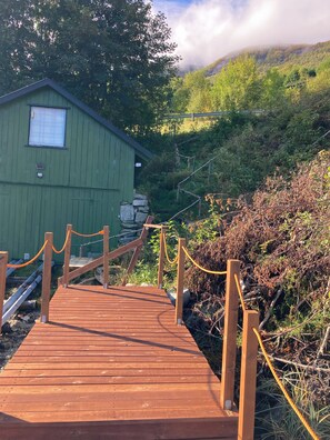 A wooden deck leading to a green building with a window, surrounded by natural vegetation and a mountainous backdrop. - Cozy vacation rental on the Langfjord in Vistdal with panoramic views (Møre og Romsdal)
