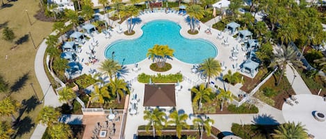 An aerial view of a resort with a central swimming pool, surrounded by lounge areas with umbrellas, palm trees, and a beach area.