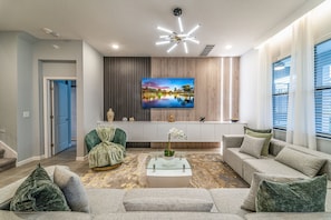 A modern living room with a glass coffee table, a sofa, and a television mounted on the wall. - Luxury 8BR Villa w/ Cinema, Game Room, Pool (Orlando)