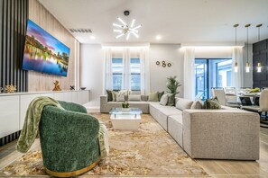 A modern living room with a large flat-screen TV, a sectional sofa, a glass coffee table, and a decorative plant. - Luxury 8BR Villa w/ Cinema, Game Room, Pool (Orlando)