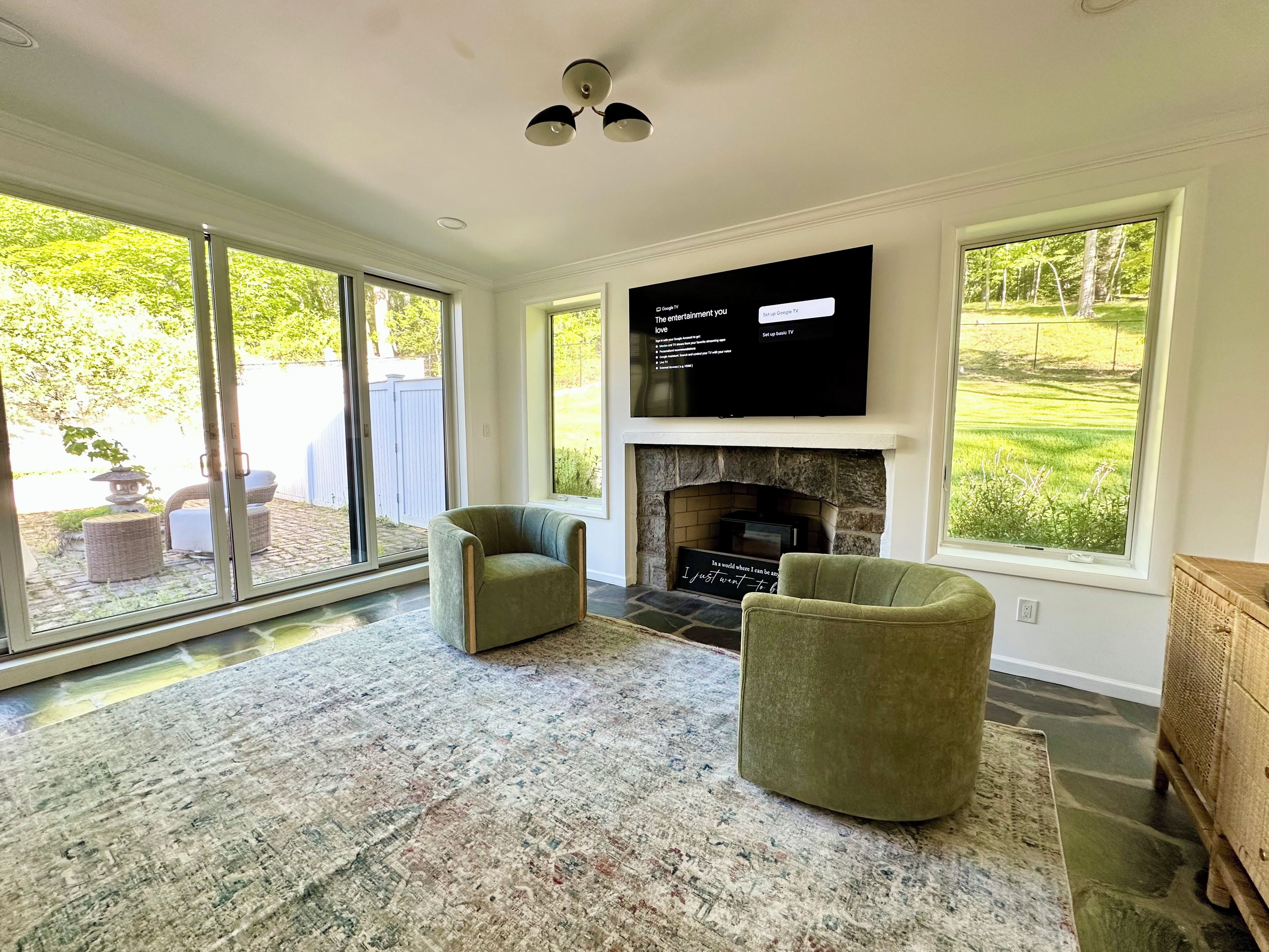A living room with a fireplace, two armchairs, a flat-screen TV, and large windows overlooking a garden.