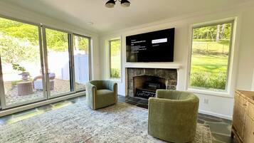 A living room with a fireplace, two armchairs, a flat-screen TV, and large windows overlooking a garden.