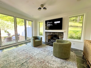 Una sala de estar con chimenea, dos sillones, un televisor de pantalla plana y grandes ventanas con vistas a un jardín.