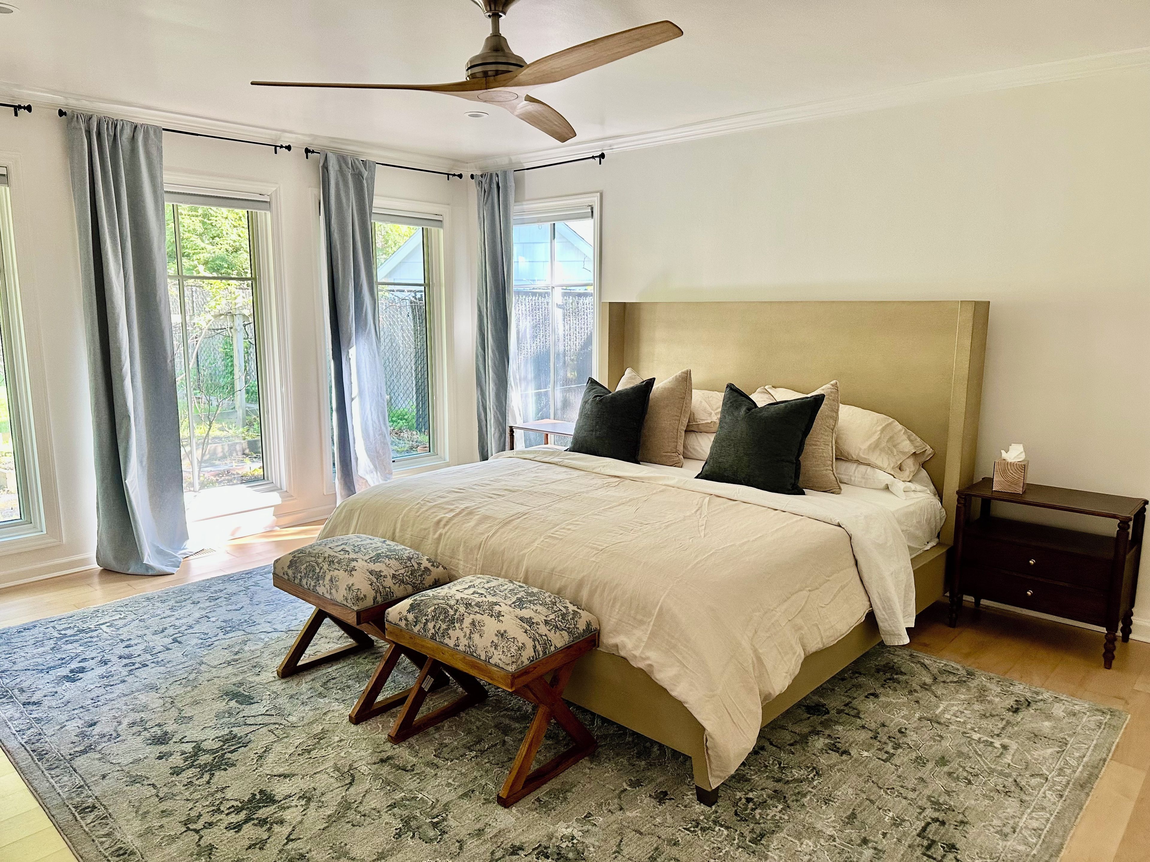 Une chambre à coucher comprenant un lit, une table de chevet, un banc, un ventilateur de plafond et de grandes fenêtres.