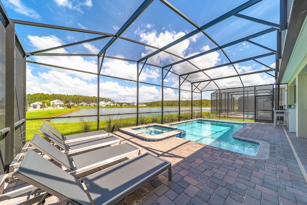 A covered outdoor pool area with lounge chairs, a hot tub, and a view of a lake and greenery. - Storey Lake 9BR Home w/ Pool & Epic Game Room (Kissimmee)