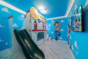 Une pièce avec un toboggan, un espace de jeux agrémenté d’un mur décoré sur le thème d’un personnage, et un téléviseur.