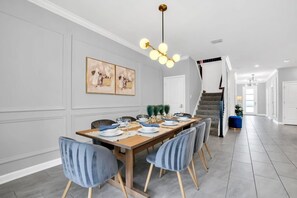 A dining area with a long wooden table set for six, surrounded by six chairs with wooden legs. There is a chandelier above the table, and the walls are painted in a light grey color. - Storey Lake 9BR Villa w Pool, Theater, Game Room (Kissimmee)