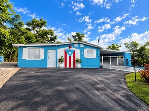 A single-story building with a blue exterior, a white door, and a garage with a white gate. - Cozy 1-bedroom apartment in serene Aguas Buenas (Aguas Buenas)