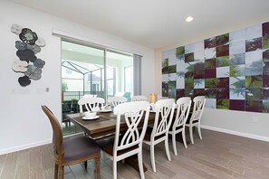 A dining area with a wooden table and white chairs, a large window, and a wall with a mosaic of nature images. - 9BR Disney Home w/ Pool, Spa & Theater Room (Kissimmee)