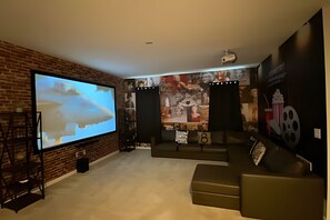 A cozy living room with a large flat-screen TV, a brick accent wall, a sectional sofa, and a projector screen. - 9BR Disney Home w/ Pool, Spa & Theater Room (Kissimmee)