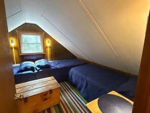 A small, cozy attic bedroom with two beds, a wooden chest, and a window. - Carrabassett Cabin | Minutes to Sugarloaf (Carrabassett Valley)