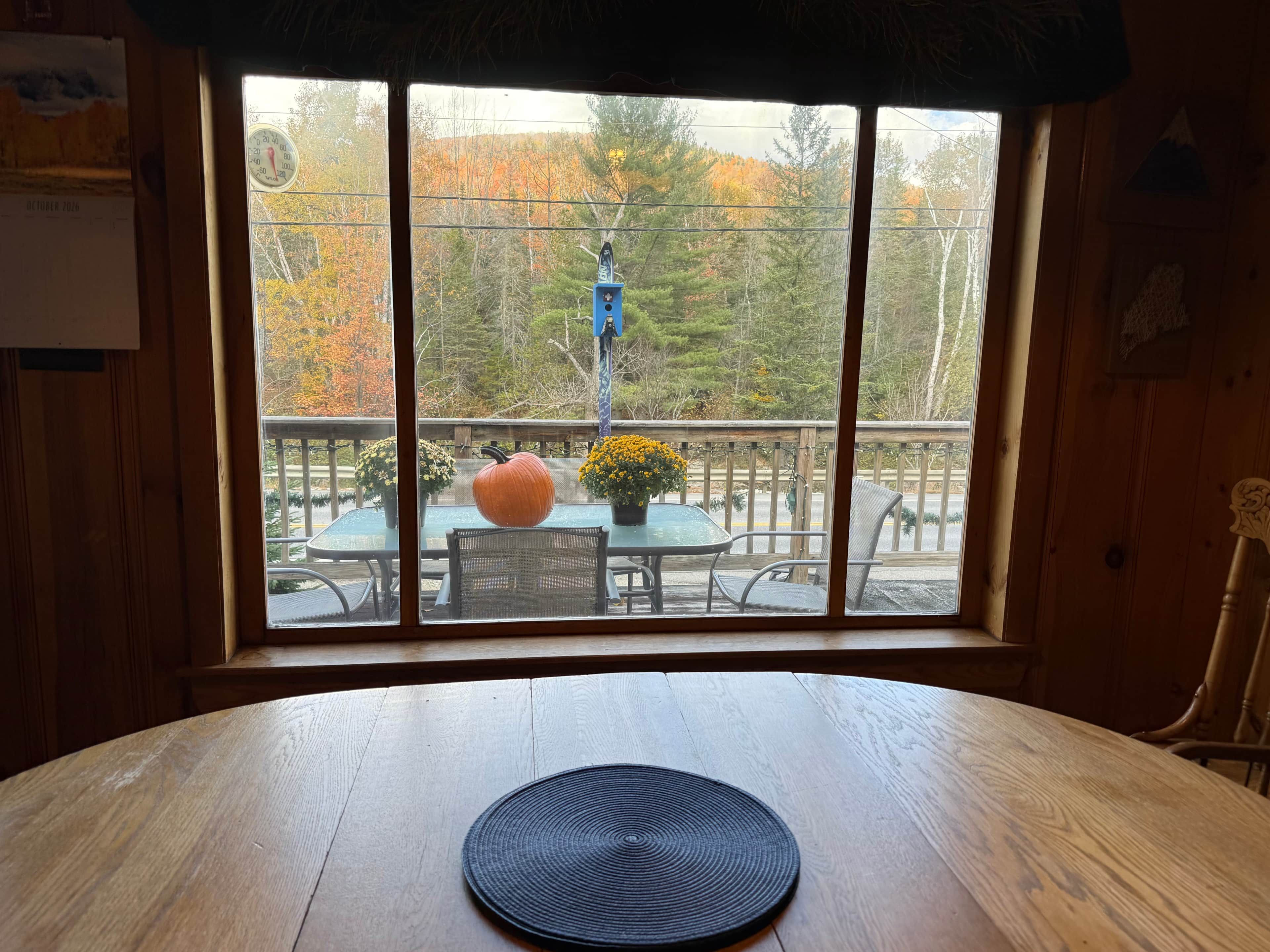 A wooden table with a black placemat, a pumpkin, and a flower vase on a balcony with a view of trees and a blue hanging ornament.