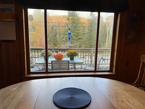 A wooden table with a black placemat, a pumpkin, and a flower vase on a balcony with a view of trees and a blue hanging ornament. - Carrabassett Cabin | Minutes to Sugarloaf (Carrabassett Valley)