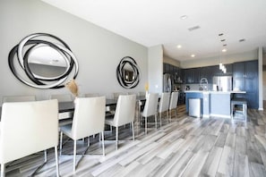 A modern kitchen with dark cabinets, a central island, and a dining area with white chairs and a glass table. - 12br Villa w/ Pool Solterra Resort Orlando (Davenport)