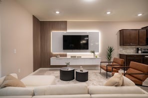 A modern living room with a sofa, two black side tables, a TV mounted on a white wall, and a kitchen area with wooden cabinets. - 10br Windsor Cay Resort Home + Private Pool (Clermont)