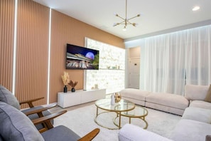 A modern living room with a sofa, a coffee table, a TV mounted on the wall, and a window with sheer curtains. - 12br Mansion w/ Private Pool 20min to Disney (Kissimmee)
