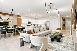 A modern living room with a sofa, chairs, a coffee table, and a dining area with a table and chairs. - 10br Themed Villa + Pool Theater & Pizza Oven (Clermont)