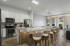 A modern kitchen with white cabinetry, stainless steel appliances, a granite countertop island with bar stools, and a view of the outdoors. - Solterra 12 BDR 10 Suites 29 Guests Pool/spa (Davenport)