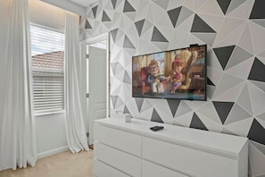 A modern living room with a flat-screen TV mounted on a geometric-patterned wall, a white entertainment center, and a window with white curtains. - Solterra 12 BDR 10 Suites 29 Guests Pool/spa (Davenport)