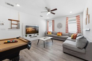 Una sala de estar con mesa de billar, sofá gris, mesa de centro y ventilador de techo.