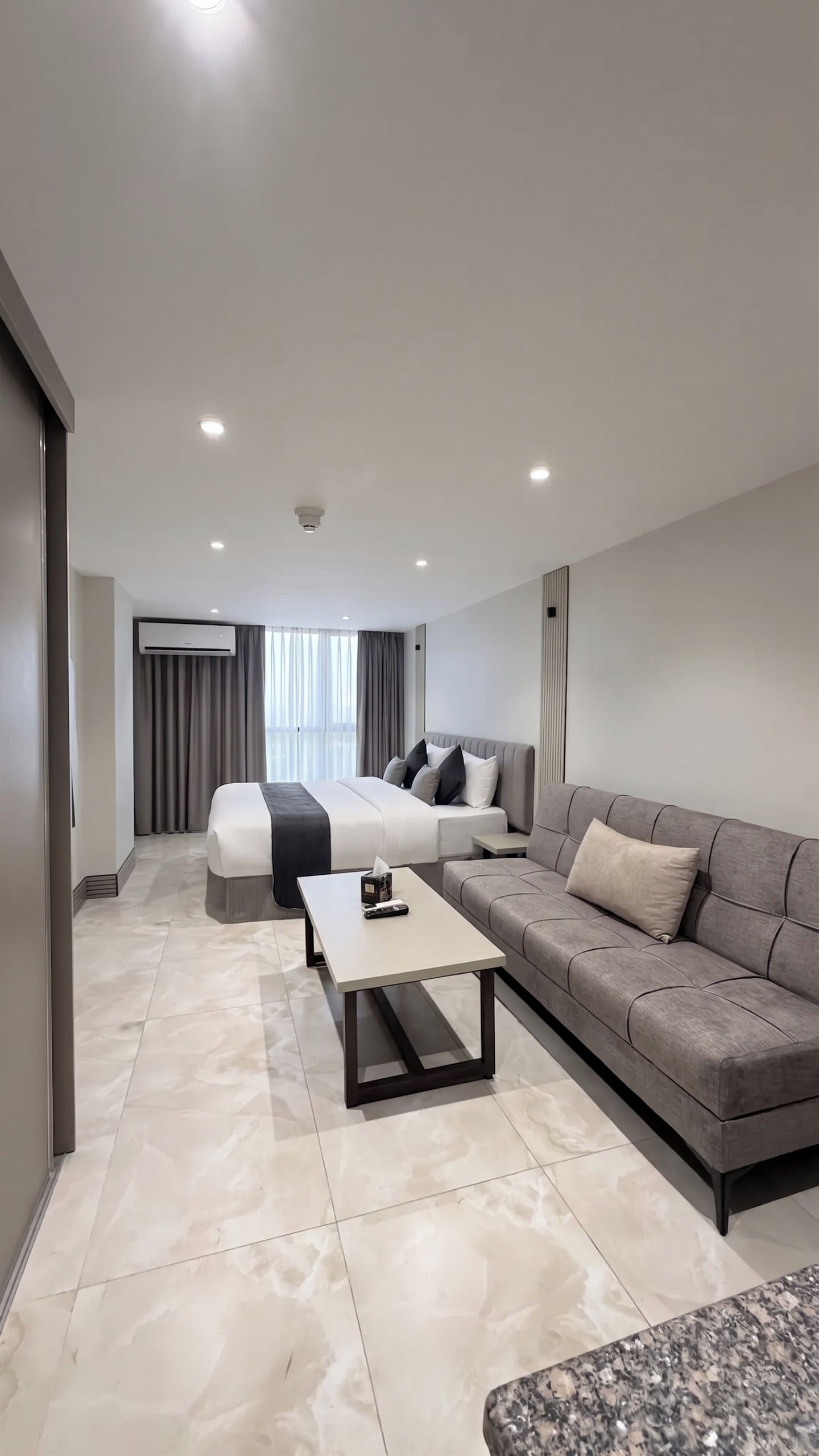 A modern hotel room with a bed, a sofa, a coffee table, and a large window with curtains.