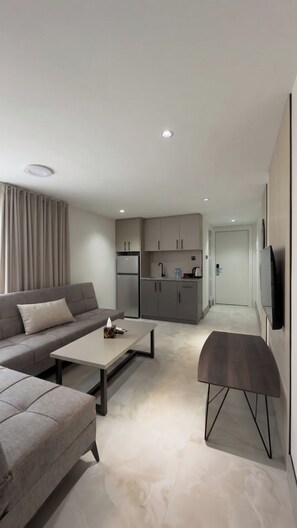 Una sala moderna con un sofá, una mesita de centro y un banco. Hay una zona de cocina con armarios y un refrigerador. Un televisor está montado en la pared.