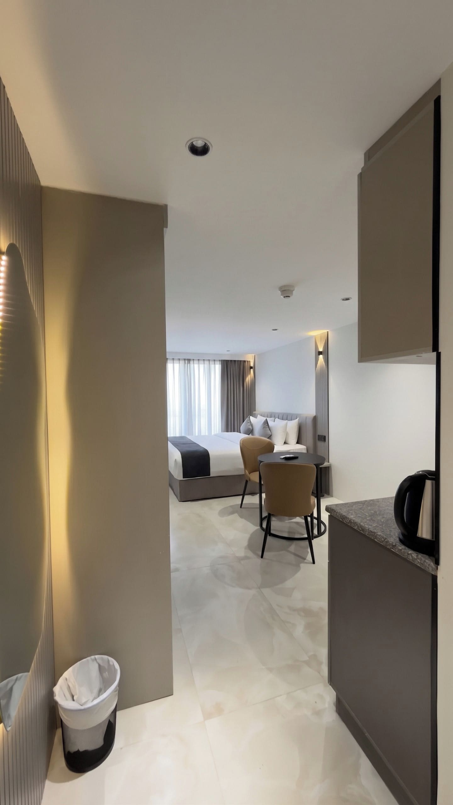 A modern hotel room with a bed, a small seating area, and a kitchenette.