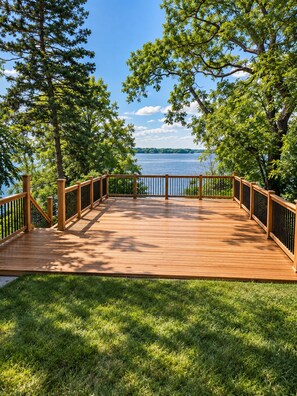 A wooden deck with a railing, surrounded by grass and trees, overlooking a body of water. - Private Lake Access, Renovated Home, Walk Downtown (Beaver Dam)