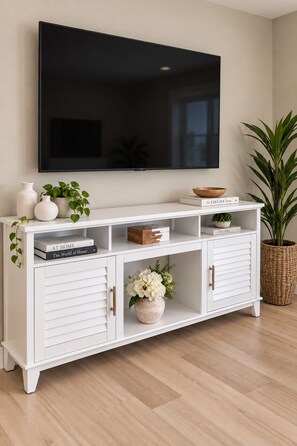 A white TV stand with a flat-screen TV mounted above it, a small wooden bowl, a book titled 'The KINFOLK Home,' and a potted plant on the right side. - Private Lake Access, Renovated Home, Walk Downtown (Beaver Dam)