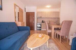 Een knus woongedeelte met een blauwe bank, een houten salontafel, een eettafel met stoelen en een kitchenette op de achtergrond.