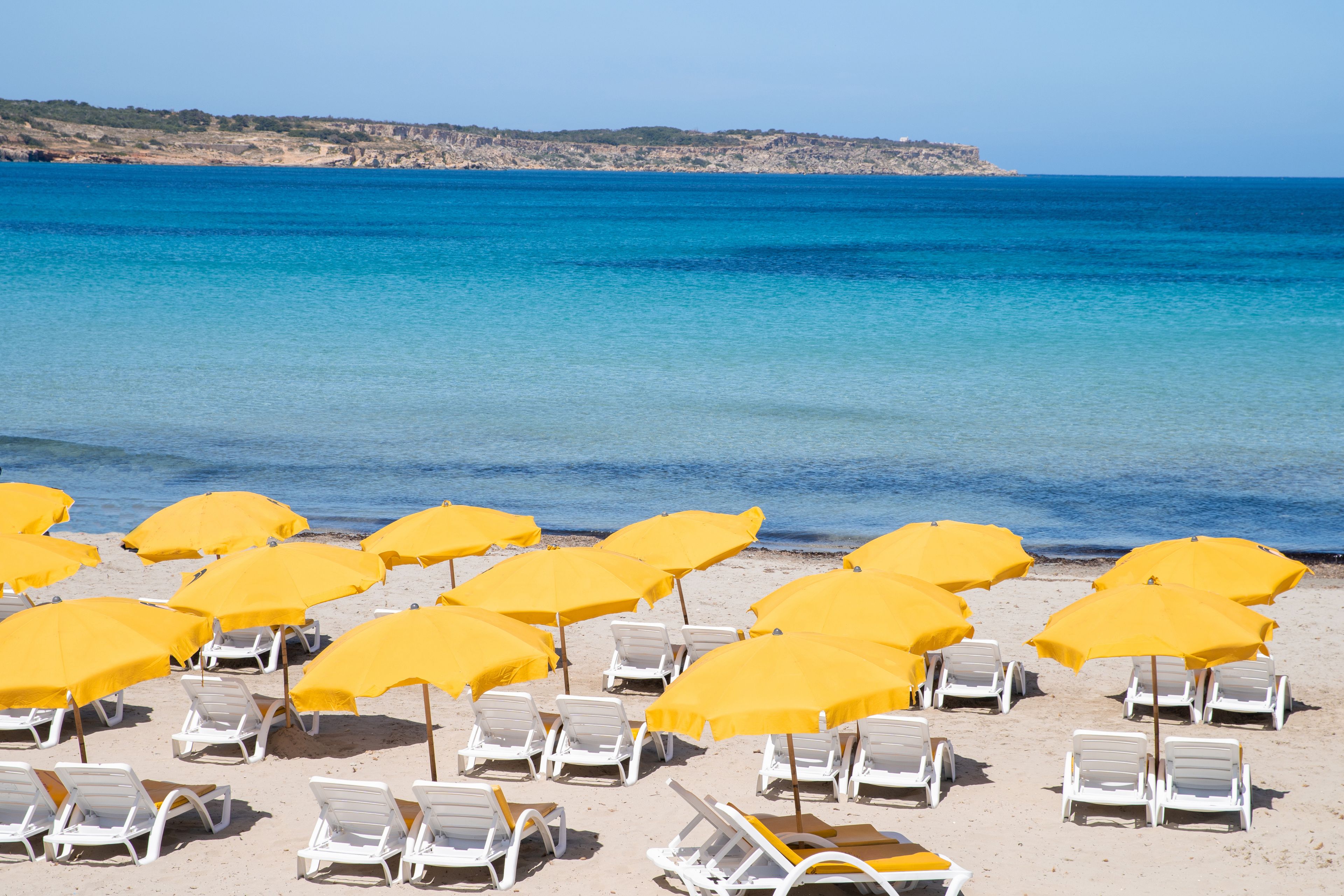 A beach with rows of white lounge chairs and yellow umbrellas, a clear blue sea, and a rocky coastline in the background.
