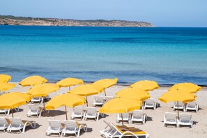 Ein Strand mit Reihen weißer Liegestühle und gelber Sonnenschirme, ein klares blaues Meer und eine felsige Küstenlinie im Hintergrund.