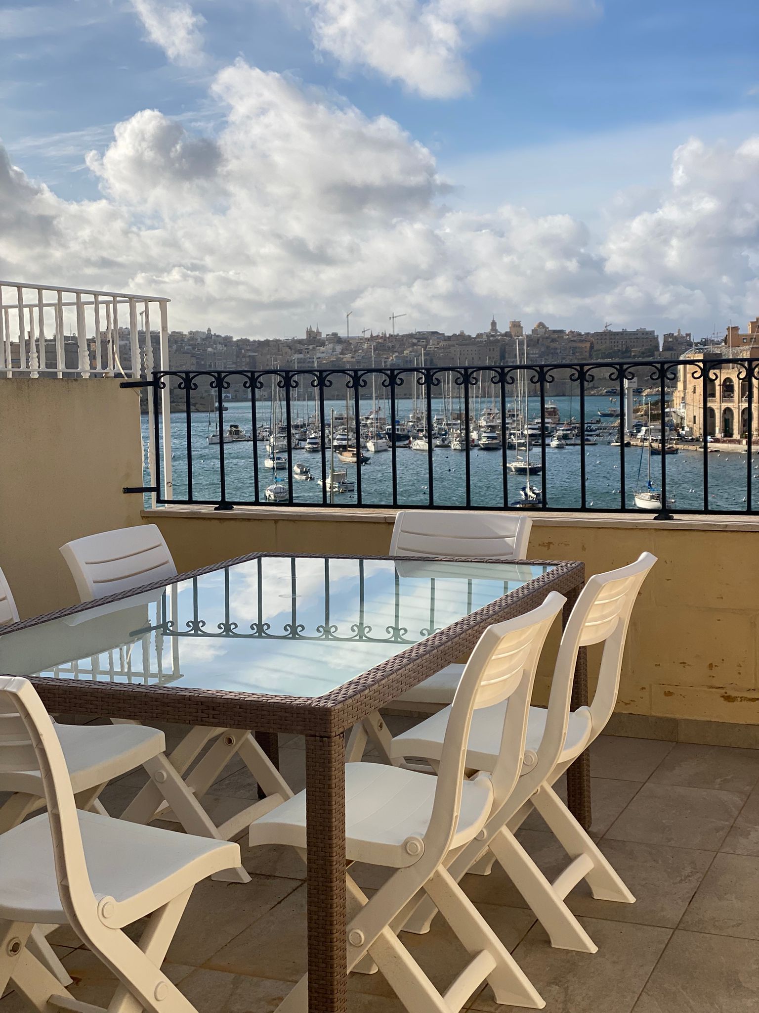 A balcony with a glass table and white chairs, overlooking a marina with boats and buildings.