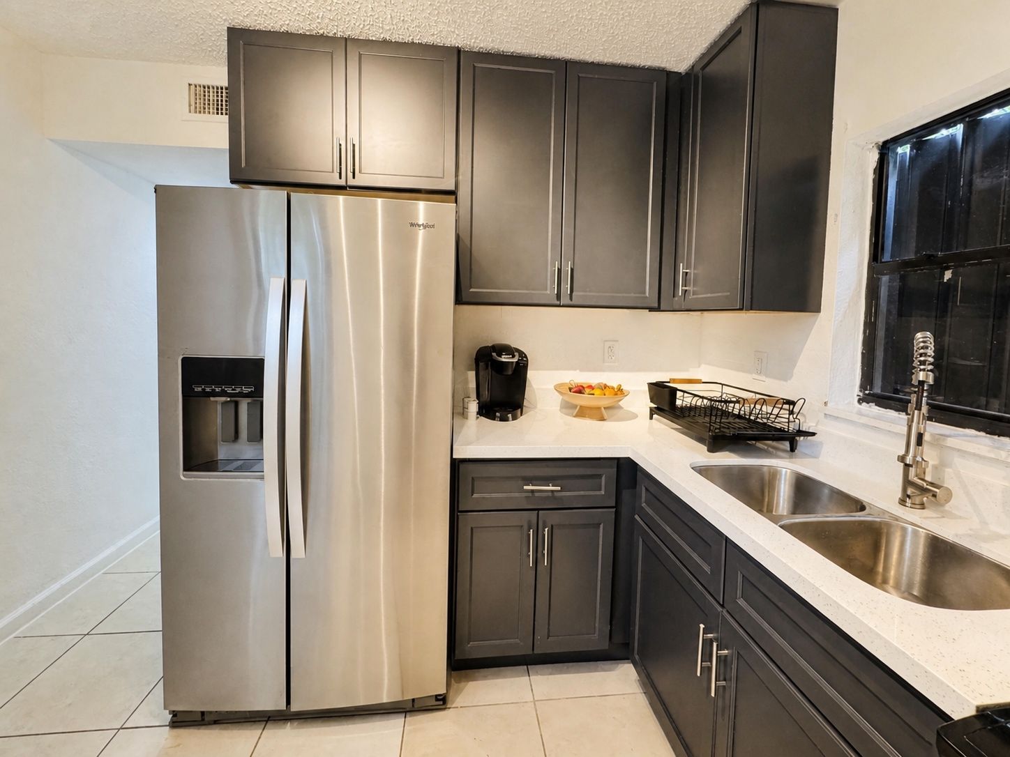 Una cocina con gabinetes oscuros, un refrigerador de acero inoxidable, una cafetera y un lavamanos.