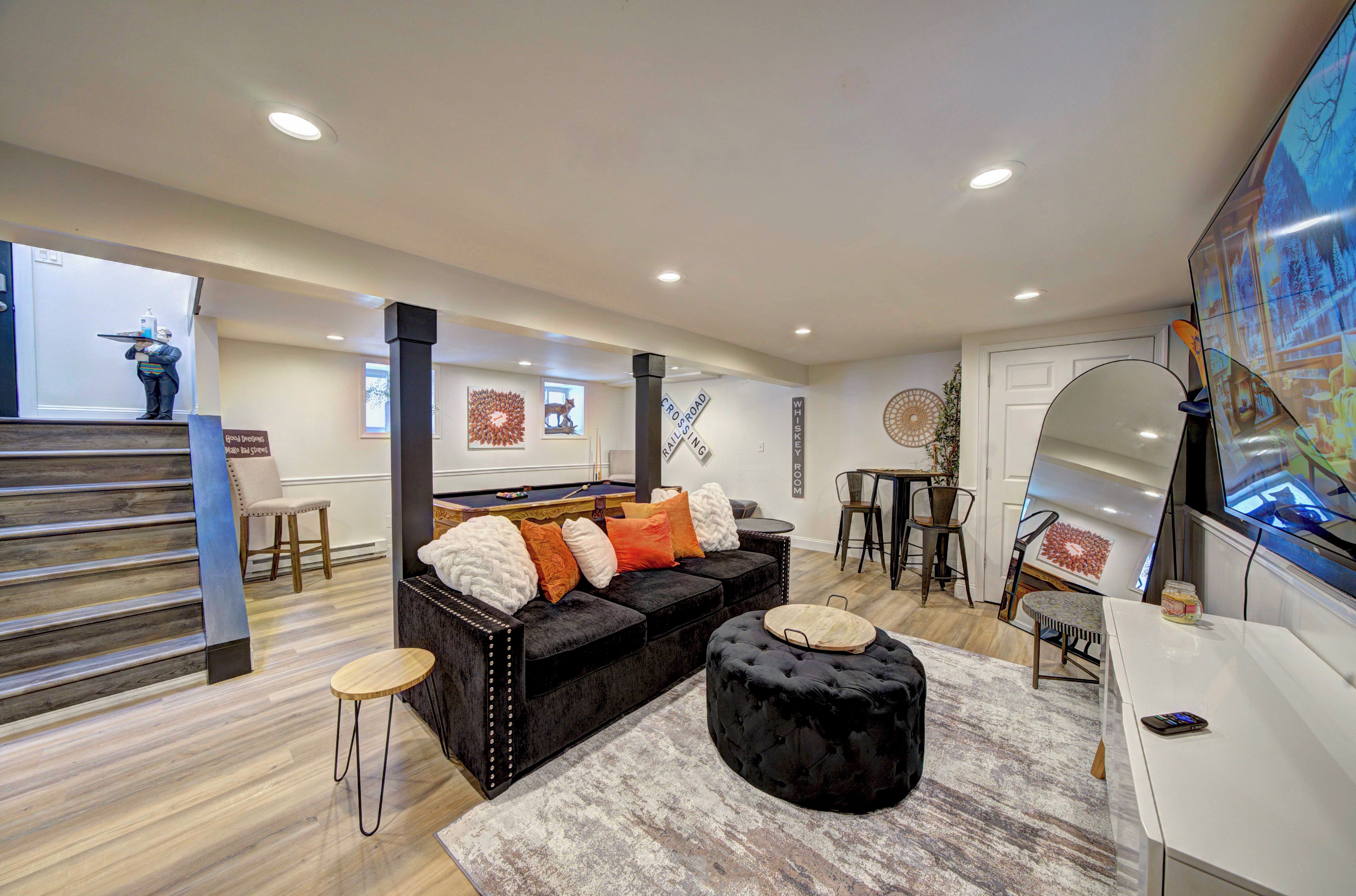A modern living room with a black sofa, a round ottoman, and a pool table.