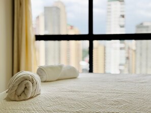 A neatly made bed with a rolled towel, a window with a black frame, and a view of buildings outside. - Apartment 3S across from the Shopping Center next to Vaca Brava Park (Goiânia)