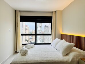 A hotel room with a bed, a view of tall buildings, and a window with curtains.