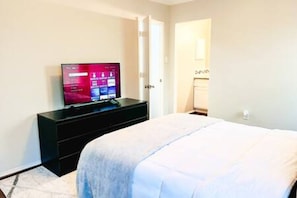 A hotel room with a bed, a TV on a stand, a door, and a light switch on the wall. - Backyard + Office + Clean + Comfy Beds (Fort Worth)