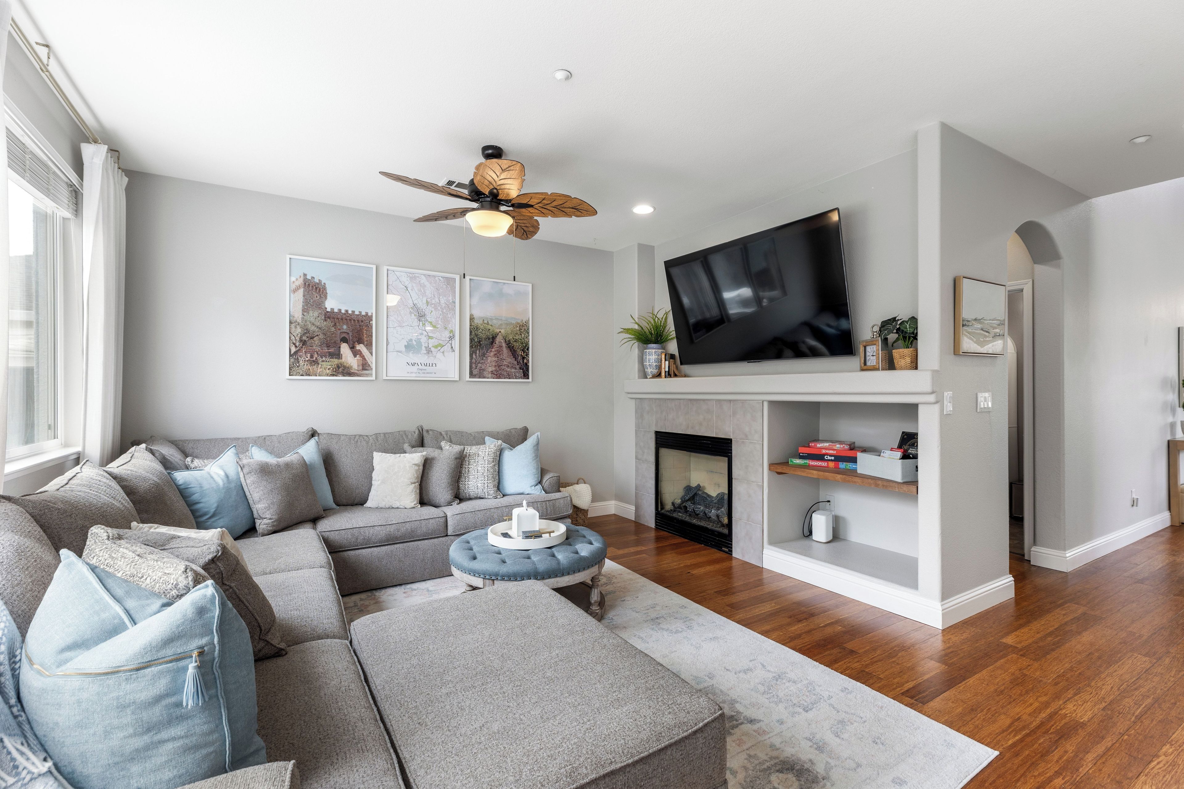 A modern living room with a sectional sofa, a flat-screen TV mounted above a fireplace, and a ceiling fan.