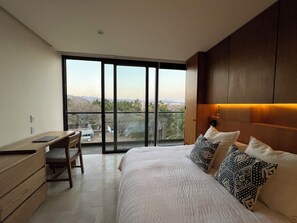 A bedroom with a large bed, a desk, and a chair. There is a balcony with a view of palm trees and buildings. - BEAUTIFUL APARTMENT BY THE BEACH   (Cruz de Huanacaxtle)
