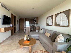 A modern living room with a sofa, coffee table, and a TV. There is a kitchen in the background with cabinets and a refrigerator. - BEAUTIFUL BEACHFRONT APARTMENT (Cruz de Huanacaxtle)